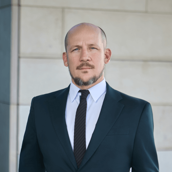 A man in a dark suit, white shirt, and black tie stands in front of a light stone wall, looking directly at the camera with a serious expression.