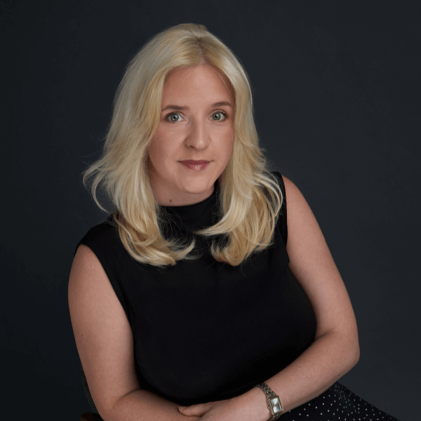 A woman with long blonde hair wearing a sleeveless black top sits against a dark background, looking at the camera with a neutral expression. She has light skin and is wearing a wristwatch.
