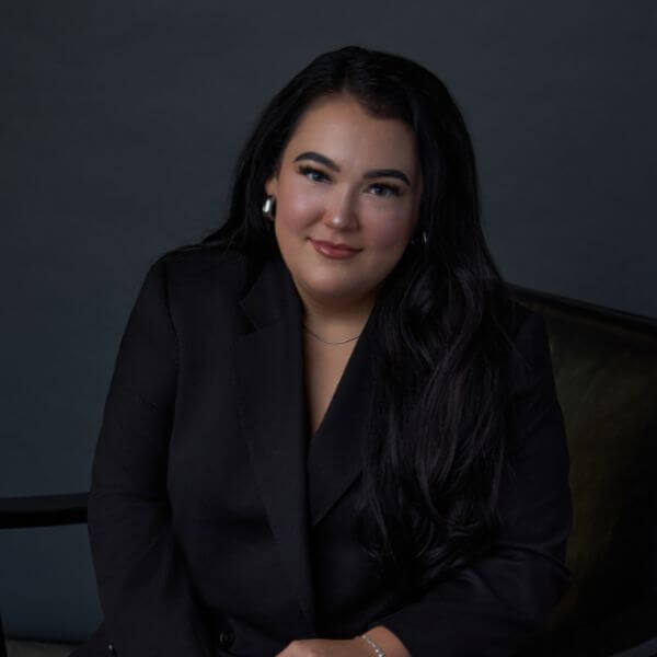 A woman with long dark hair, wearing a black blazer and silver earrings, sits on a dark chair against a dark grey background, smiling gently at the camera.