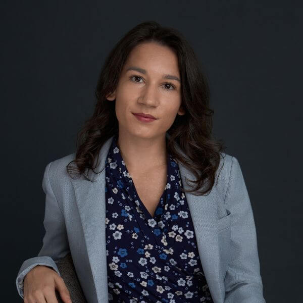 A woman with wavy brown hair wearing a light blue blazer and a navy blue floral blouse sits against a dark background, looking at the camera with a neutral expression.