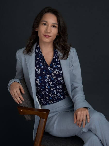 A person with long dark hair sits on a chair against a dark background, wearing a light blue suit and a navy blue floral blouse, looking confidently at the camera.