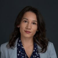 A person with long brown hair wearing a light blue blazer over a blue floral blouse, posing against a dark background and looking at the camera with a neutral expression.